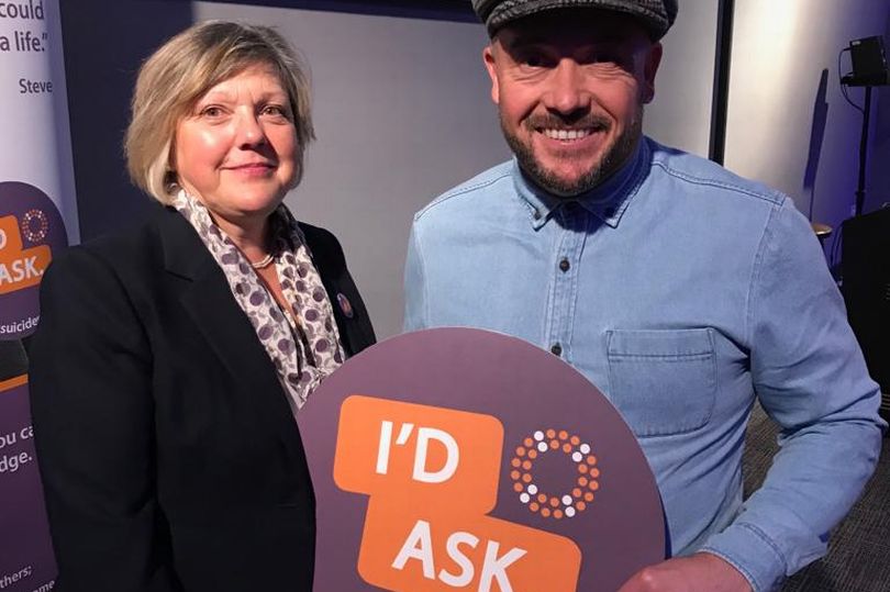 A white woman and white man holding a purple and orange I'd Ask sign