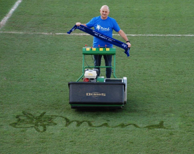 Man in a blue CPSL Mind t shirt holding a whoever you support we support you scarf on a football pitch in front of a lawn mower with the Mind logo mown into the grass