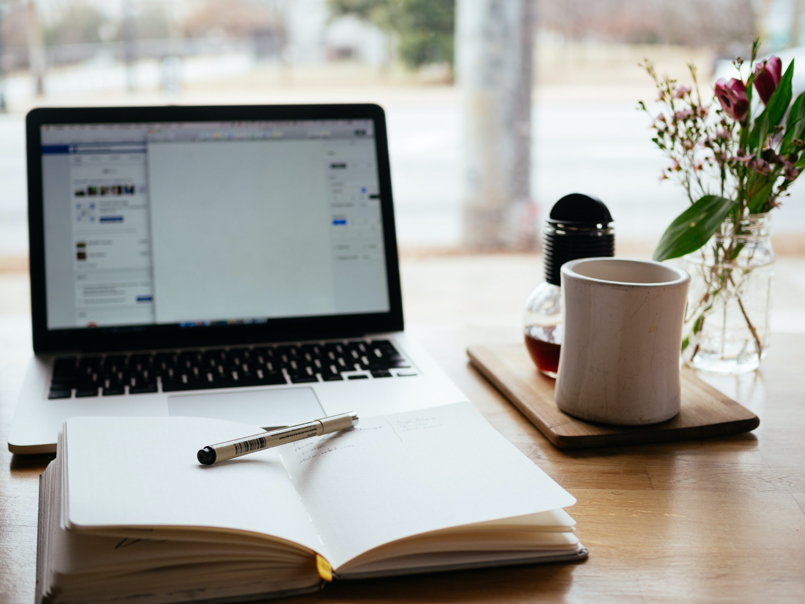 Laptop on desk with a notepad open with pen on it. A coffee cup and coffee maker is next to flowers in a vase