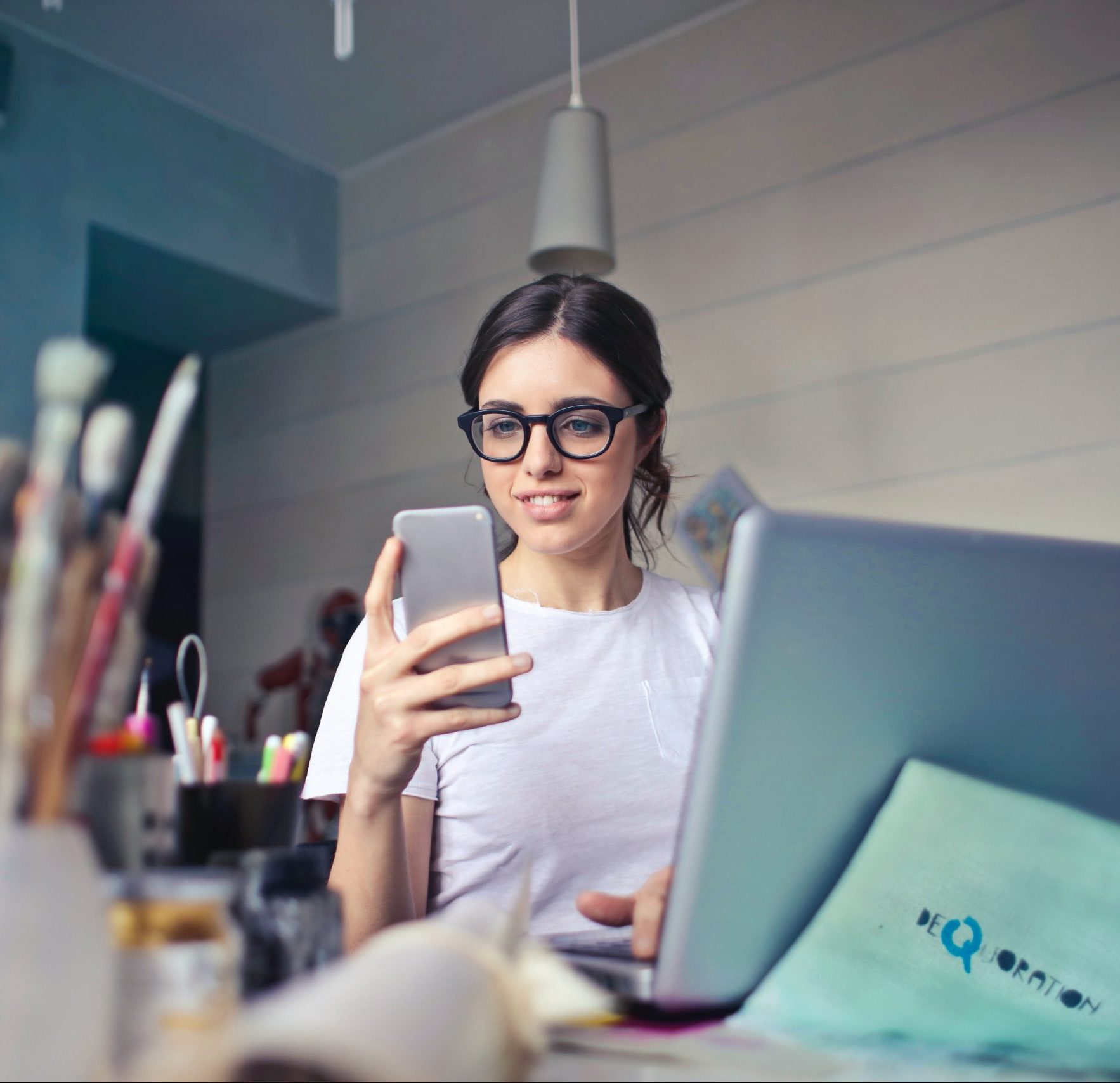 dark haired girl with glasses sat at a desk with her laptop, looking at her phone