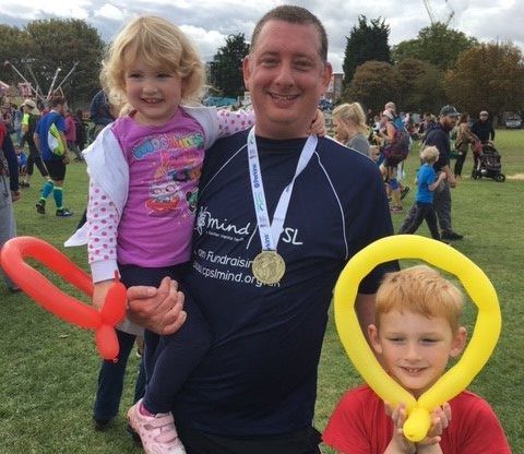 A man with a medal and CPSL Mind tshirt holding a little girl, with a little boy holding a yellow long balloon around his face