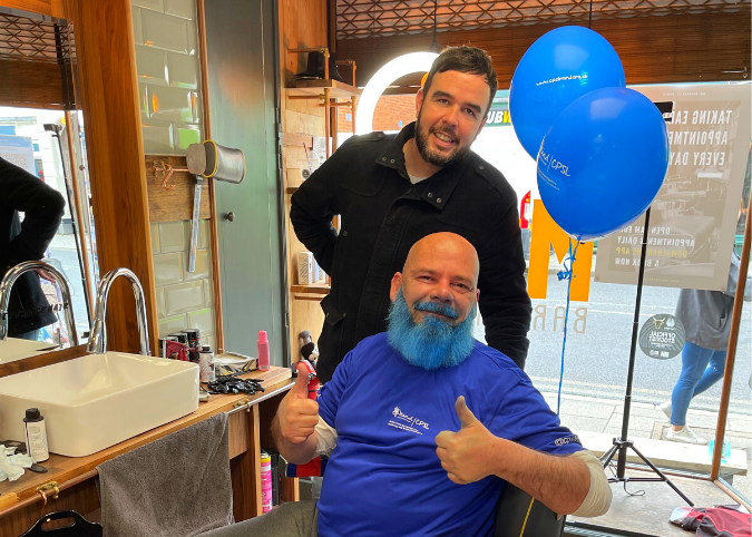 Two men in a barbers one with his beard dyed blue