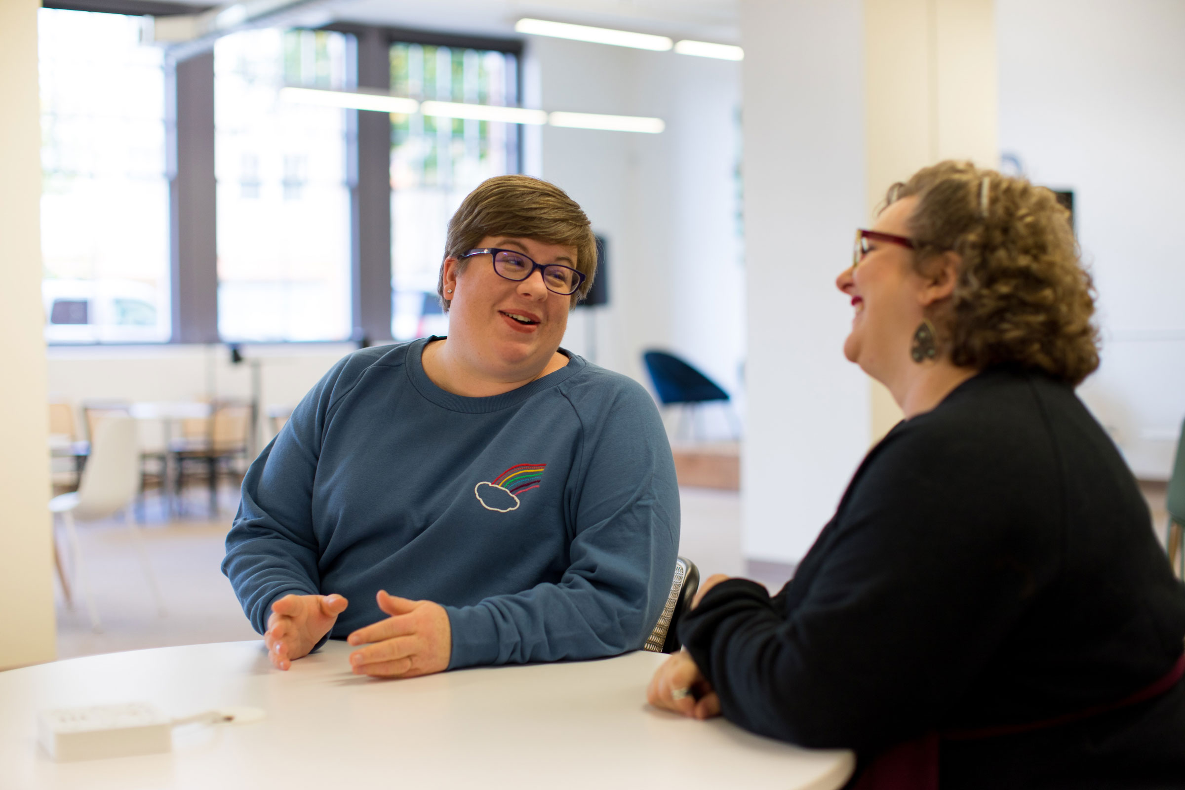 Two people with glasses sat in a white room at a table smiling