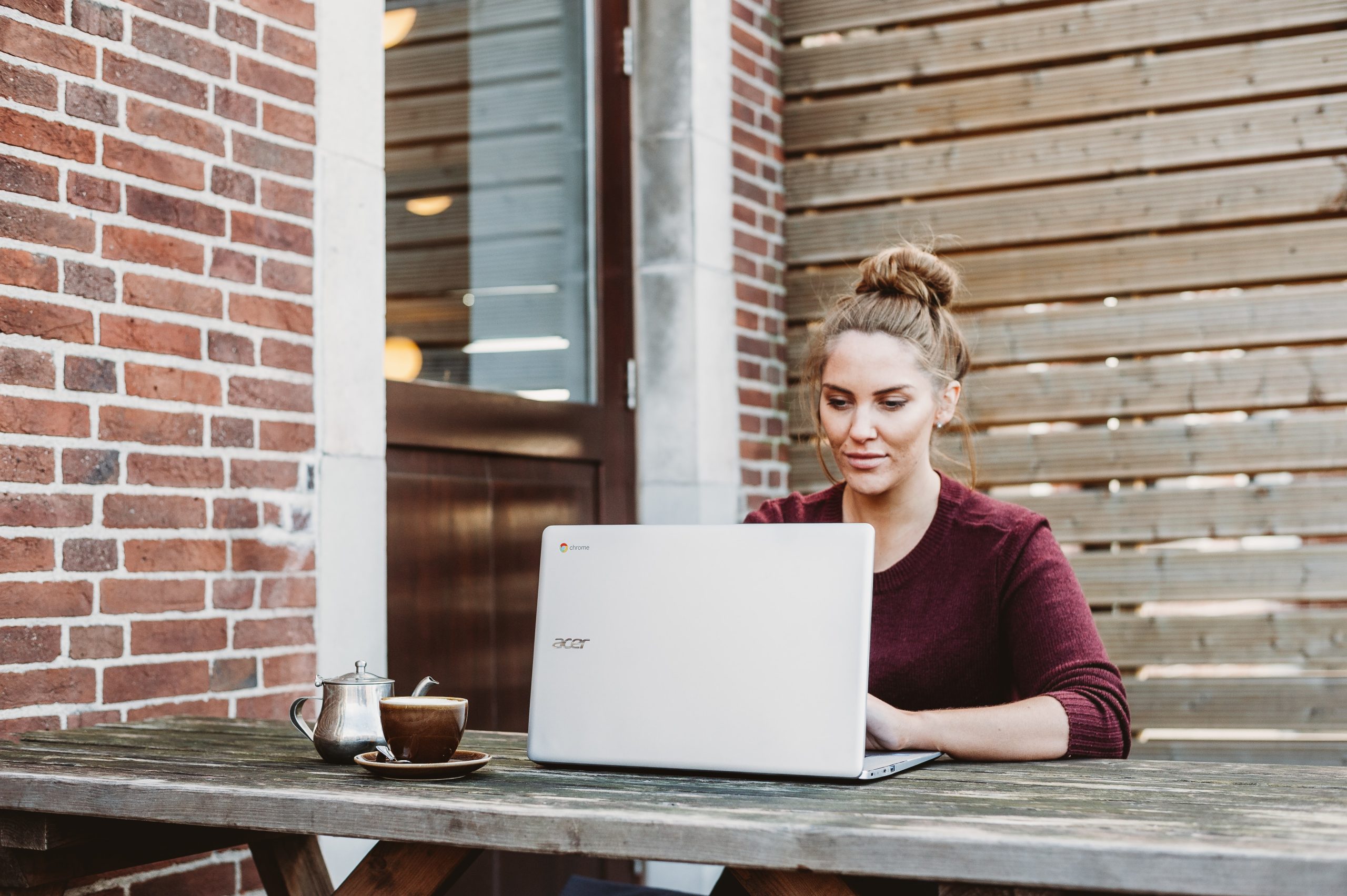 Women in red jumper and hair tied up working on laptop outside on table with coffee cup