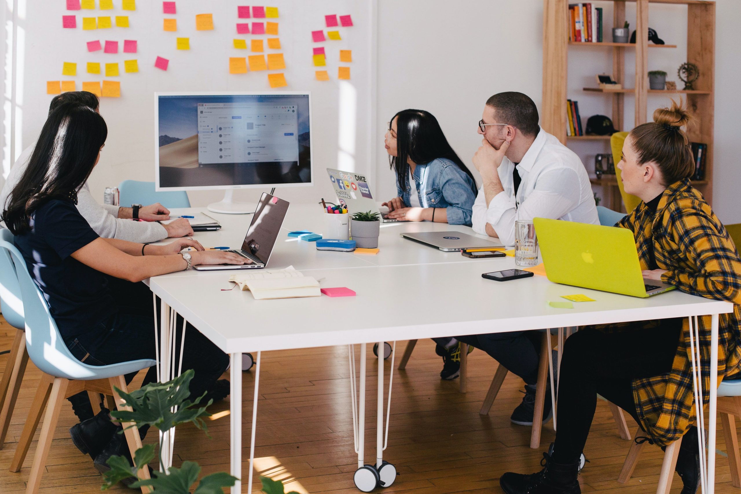 Group of young office workings on laptop sitting around a desk looking at digital screen with sticky notes around it